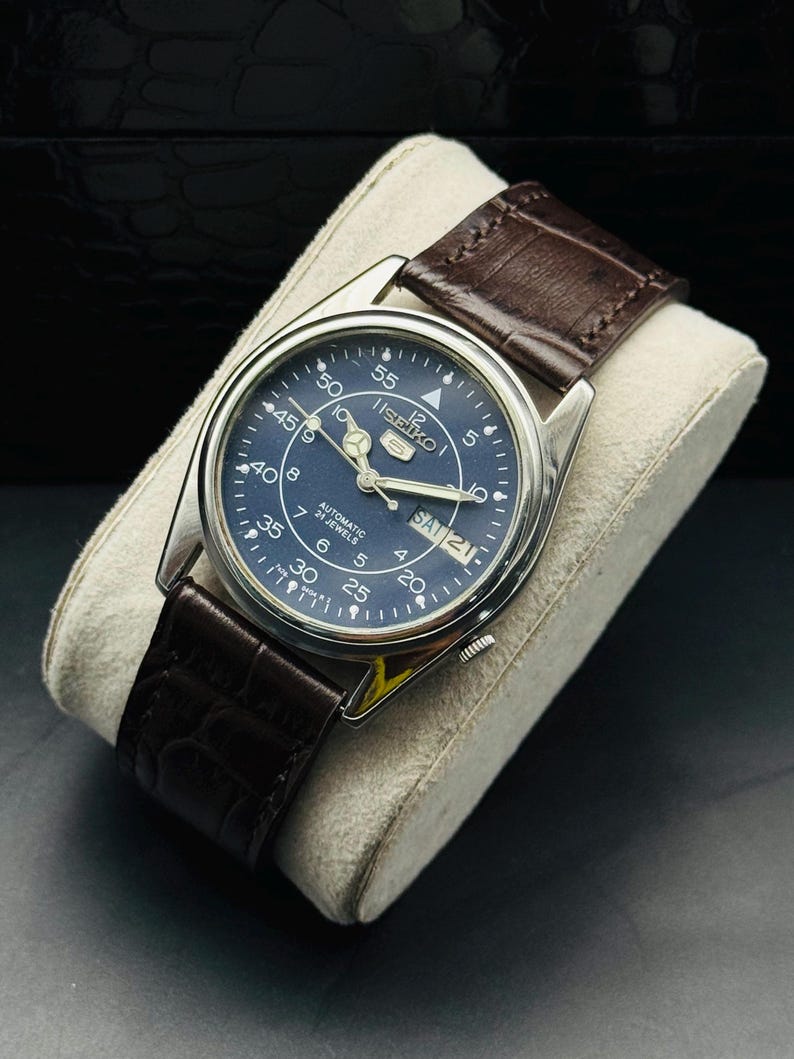 May include: A silver-toned Seiko automatic watch with a dark blue face and brown leather strap. The watch face displays the day and date, with white numerals and hands. The watch is displayed on a cream-colored cushion.