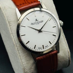 Puede incluir: Un reloj Jaeger-LeCoultre Club plateado con esfera blanca y agujas negras. Cuenta con una correa de cuero marr&oacute;n. La esfera muestra "Jaeger-LeCoultre Club" y "Swiss Made".