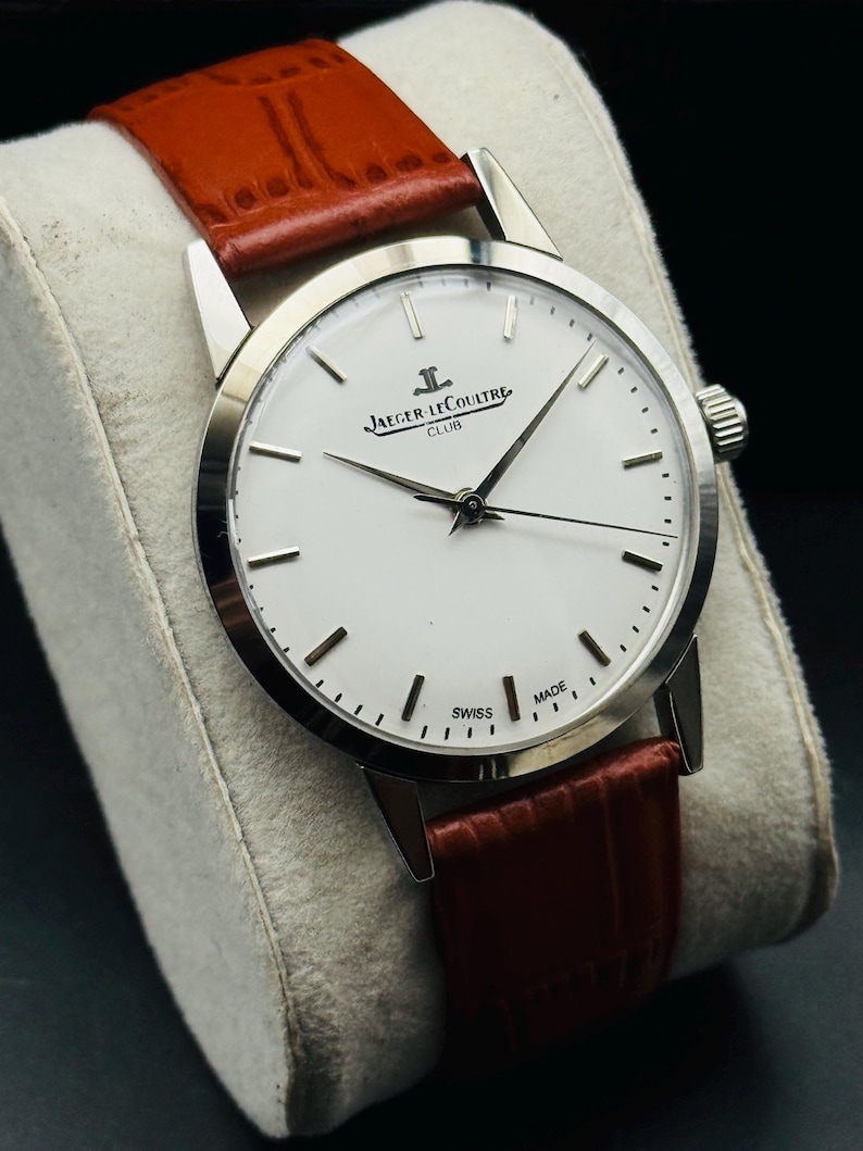 Puede incluir: Un reloj Jaeger-LeCoultre Club plateado con esfera blanca y agujas negras. Cuenta con una correa de cuero marr&oacute;n. La esfera muestra "Jaeger-LeCoultre Club" y "Swiss Made".