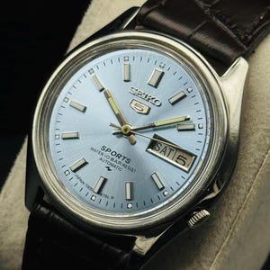 May include: A silver-toned Seiko 5 Sports automatic watch with a light blue dial and a brown leather strap. The watch face displays the day and date, with the day reading "SAT 5". The watch has a water resistance of 10 bar.