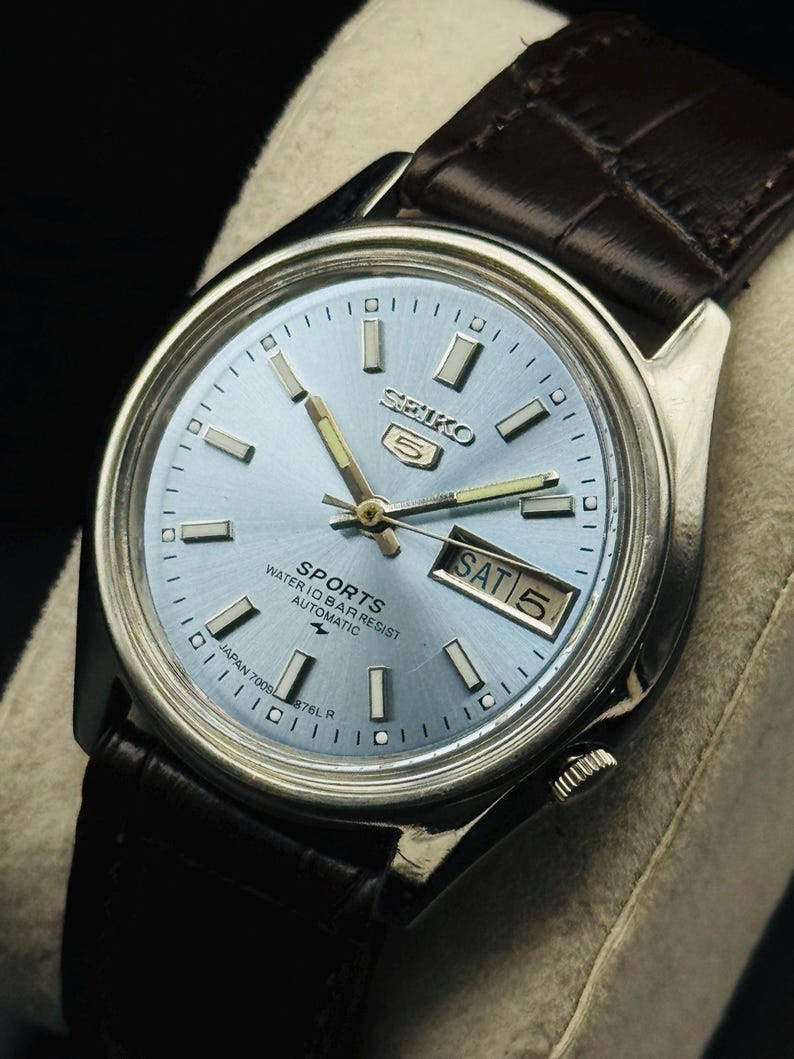 May include: A silver-toned Seiko 5 Sports automatic watch with a light blue dial and a brown leather strap. The watch face displays the day and date, with the day reading "SAT 5". The watch has a water resistance of 10 bar.