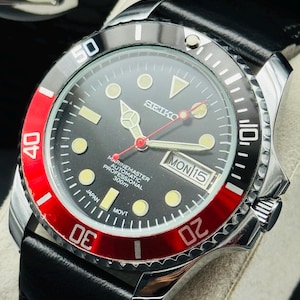 May include: A stainless steel Seiko Marinemaster automatic professional watch with a black dial, red bezel, and black leather strap. The watch face displays the day and date.