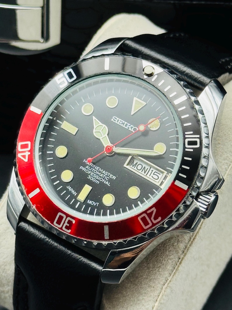 May include: A stainless steel Seiko Marinemaster automatic professional watch with a black dial, red bezel, and black leather strap. The watch face displays the day and date.