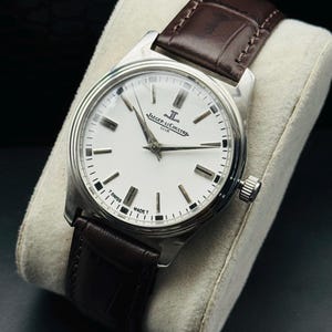 May include: A silver-toned wristwatch with a white face and black markers. The watch features the text "JAEGER-LECOULTRE CLUB" and a brown leather band. The watch is displayed on a white cushion.