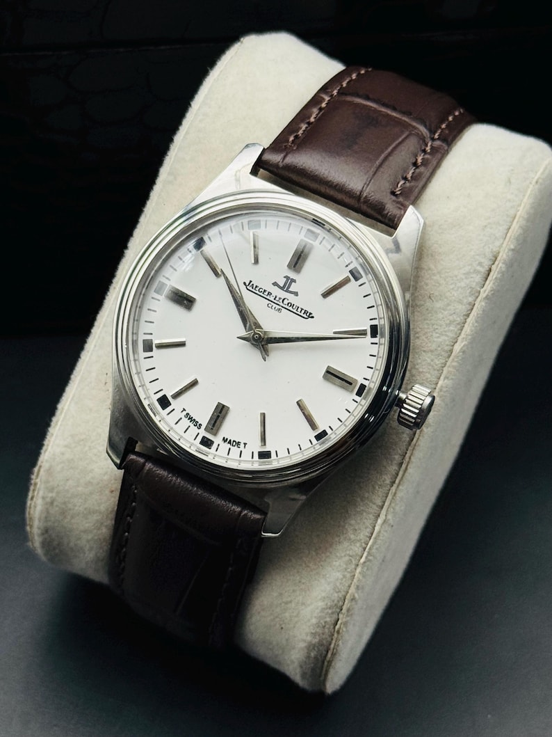 May include: A silver-toned wristwatch with a white face and black markers. The watch features the text "JAEGER-LECOULTRE CLUB" and a brown leather band. The watch is displayed on a white cushion.