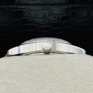 May include: A silver wristwatch with a round face and a silver band. The watch has a small, circular crown and a light-colored face. The watch is resting on a light-colored surface, with a dark, textured background.