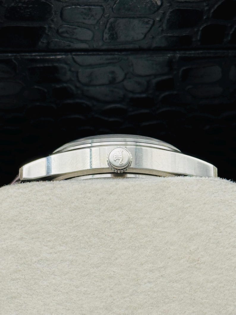 May include: A silver wristwatch with a round face and a silver band. The watch has a small, circular crown and a light-colored face. The watch is resting on a light-colored surface, with a dark, textured background.