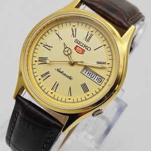 Op de afbeelding: Een goudkleurig Seiko 5 automatisch polshorloge met een crèmekleurige wijzerplaat, Romeinse cijfers en een dag-datumweergave. Het heeft een bruine leren band en wordt op een transparante standaard getoond.