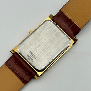 Puede incluir: Un reloj Seiko de cuarzo dorado con una esfera rectangular y una correa de cuero marr&oacute;n. La parte trasera del reloj es plateada con el logotipo y el texto de Seiko. La correa es de color marr&oacute;n oscuro.