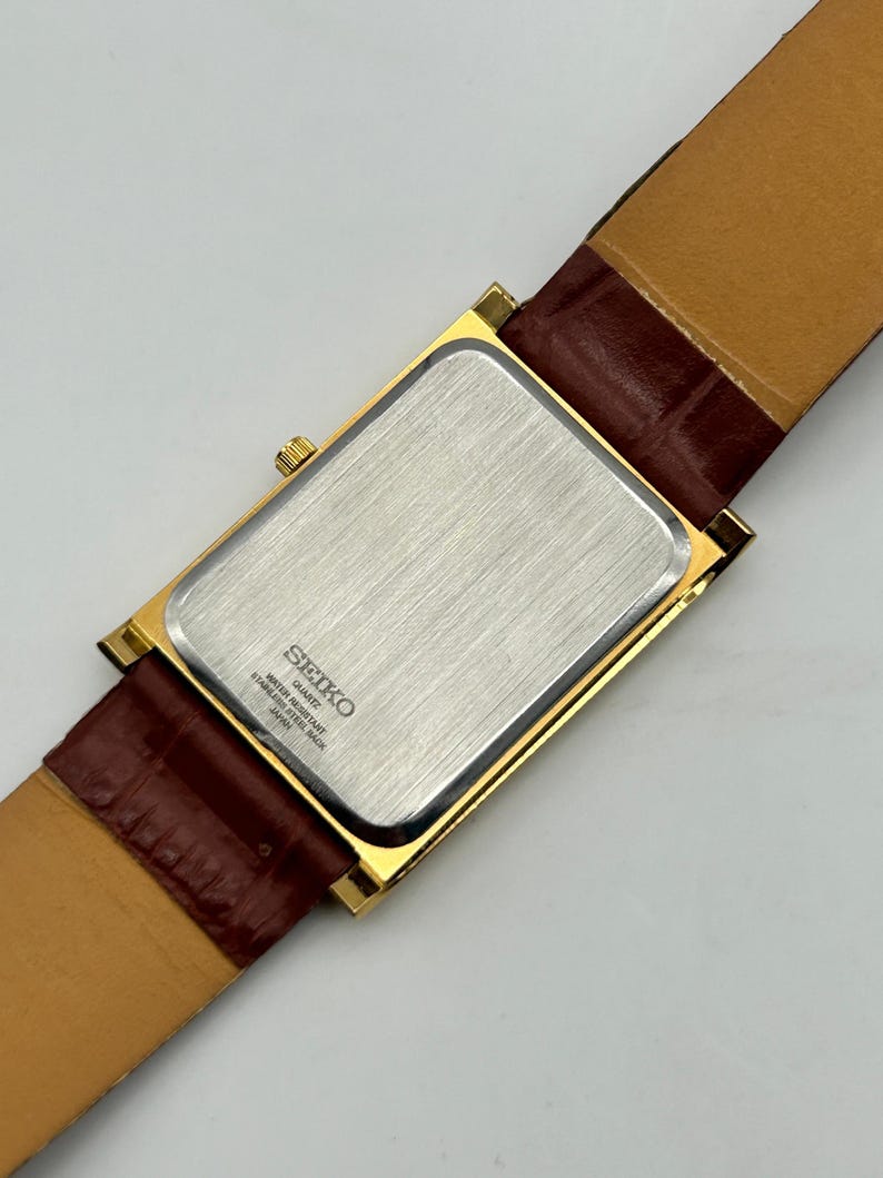 Puede incluir: Un reloj Seiko de cuarzo dorado con una esfera rectangular y una correa de cuero marr&oacute;n. La parte trasera del reloj es plateada con el logotipo y el texto de Seiko. La correa es de color marr&oacute;n oscuro.