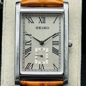 Op de afbeelding: Een rechthoekig Seiko polshorloge met een zilveren wijzerplaat, zwarte Romeinse cijfers en een kleine secondenwijzer. Het horloge heeft een roestvrijstalen kast en een bruine leren band met witte stiksels.
