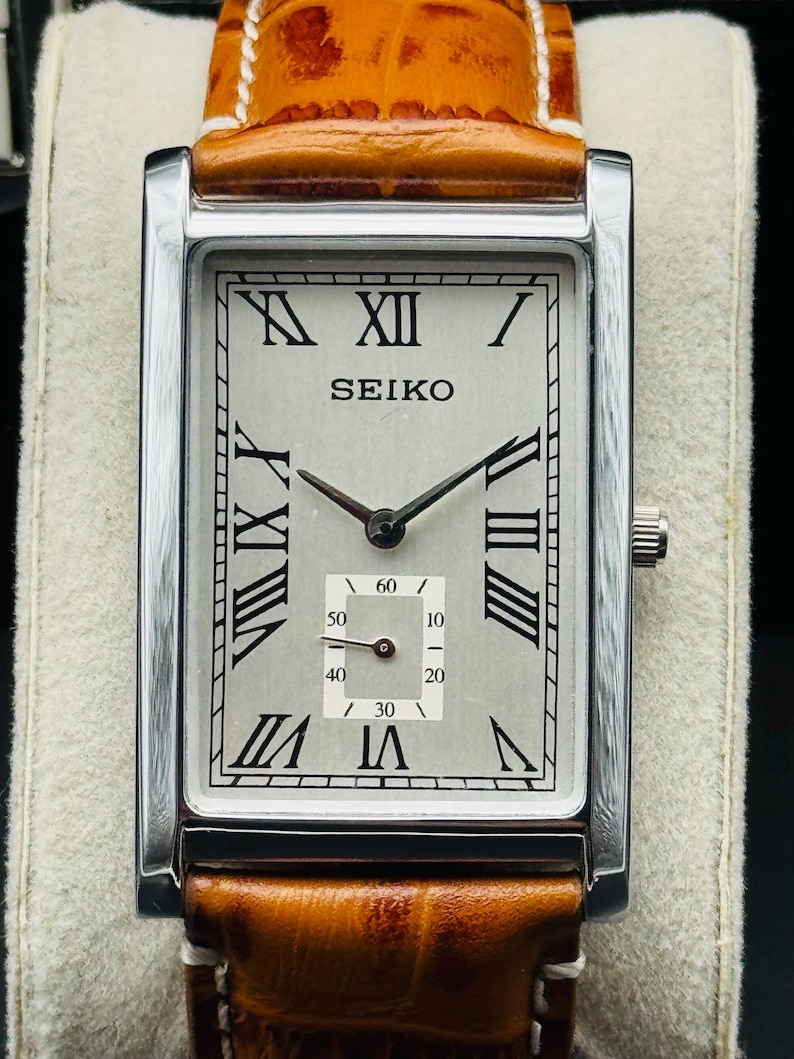 Op de afbeelding: Een rechthoekig Seiko polshorloge met een zilveren wijzerplaat, zwarte Romeinse cijfers en een kleine secondenwijzer. Het horloge heeft een roestvrijstalen kast en een bruine leren band met witte stiksels.