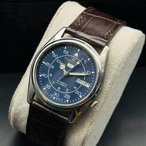May include: A silver-toned Seiko automatic wristwatch with a dark blue face and brown leather strap. The watch face displays the day and date, with white numerals and hands. The strap is a dark brown, textured leather.