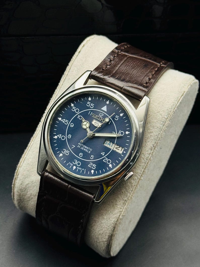 May include: A silver-toned Seiko automatic wristwatch with a dark blue face and brown leather strap. The watch face displays the day and date, with white numerals and hands. The strap is a dark brown, textured leather.
