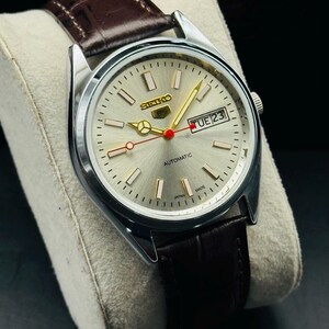 May include: A silver-toned Seiko automatic wristwatch with a light-colored face, gold hands, and a brown leather strap. The watch face displays the day and date, with the word "AUTOMATIC" printed below the center.
