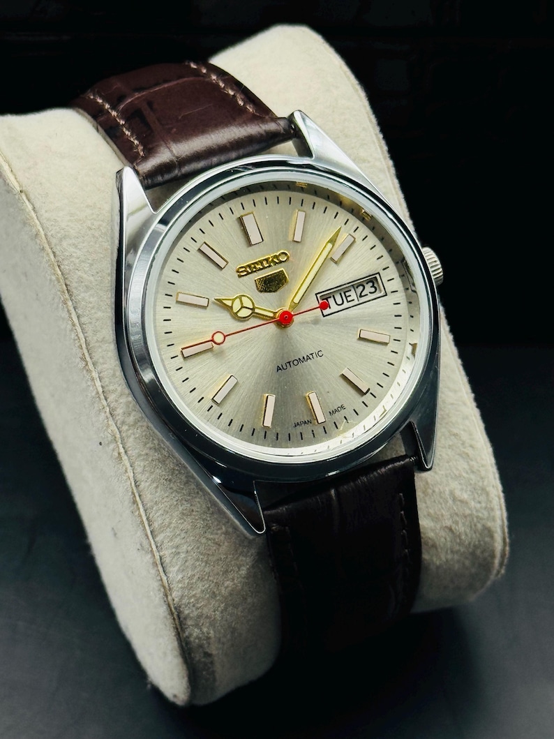 May include: A silver-toned Seiko automatic wristwatch with a light-colored face, gold hands, and a brown leather strap. The watch face displays the day and date, with the word "AUTOMATIC" printed below the center.