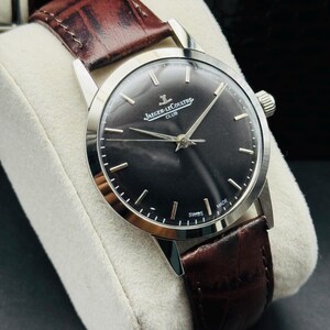 May include: A silver-toned wristwatch with a dark face and brown leather band. The watch face features silver hands and markers, with the text "JAEGER-LECOULTRE CLUB" and "SWISS MADE". The band is a rich brown color.