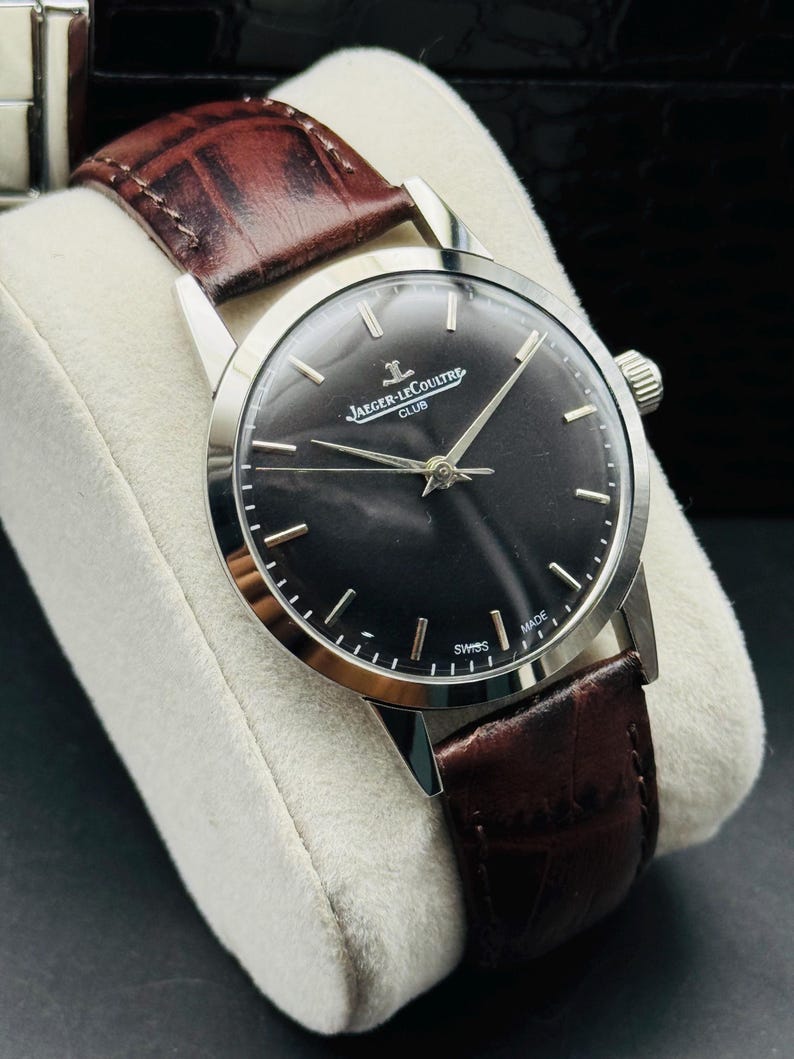 May include: A silver-toned wristwatch with a dark face and brown leather band. The watch face features silver hands and markers, with the text "JAEGER-LECOULTRE CLUB" and "SWISS MADE". The band is a rich brown color.