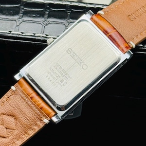Op de afbeelding: Een rechthoekig Seiko horloge met een zilverkleurige kast en een bruine leren band. De achterkant van het horloge is zichtbaar, met het Seiko-logo en tekst die aangeeft dat het waterbestendig is. De band is gestikt met witte draad.