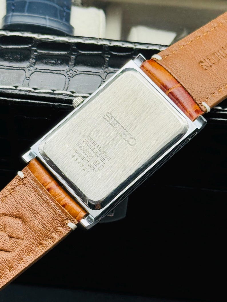 Op de afbeelding: Een rechthoekig Seiko horloge met een zilverkleurige kast en een bruine leren band. De achterkant van het horloge is zichtbaar, met het Seiko-logo en tekst die aangeeft dat het waterbestendig is. De band is gestikt met witte draad.