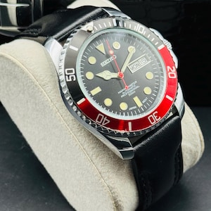 May include: A black and silver wristwatch with a black leather band. The watch face is black with luminous markers, a red second hand, and a date display. The bezel is black and red, with white numerals. The watch is labeled "SEIKO MARINE MASTER AUTOMATIC PROFESSIONAL 300m".