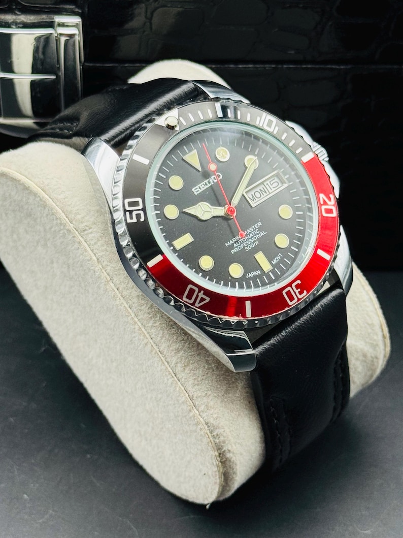 May include: A black and silver wristwatch with a black leather band. The watch face is black with luminous markers, a red second hand, and a date display. The bezel is black and red, with white numerals. The watch is labeled "SEIKO MARINE MASTER AUTOMATIC PROFESSIONAL 300m".