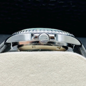 May include: A side view of a silver wristwatch with a black leather band. The watch has a light blue face and a silver bezel. The watch is resting on a white surface.