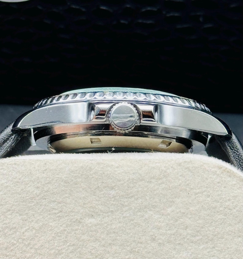 May include: A side view of a silver wristwatch with a black leather band. The watch has a light blue face and a silver bezel. The watch is resting on a white surface.