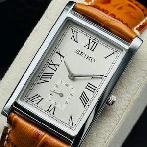 Op de afbeelding: Een rechthoekig Seiko polshorloge met een zilveren wijzerplaat en Romeinse cijfers. Het horloge heeft een bruine leren band en een zilveren kast. De wijzerplaat heeft ook een kleine seconden subdial. Het horloge wordt op een wit oppervlak weergegeven.