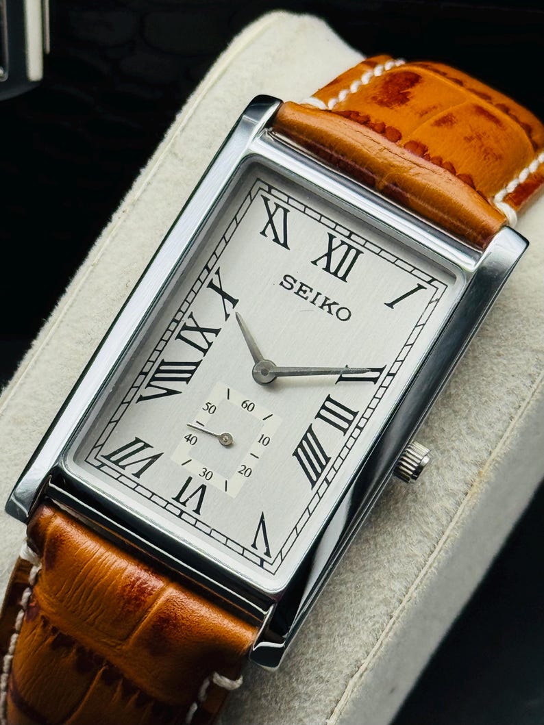 Op de afbeelding: Een rechthoekig Seiko polshorloge met een zilveren wijzerplaat en Romeinse cijfers. Het horloge heeft een bruine leren band en een zilveren kast. De wijzerplaat heeft ook een kleine seconden subdial. Het horloge wordt op een wit oppervlak weergegeven.