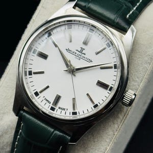 May include: A silver-toned Jaeger-LeCoultre Club watch with a white face and black markers. The watch has silver hands and a green leather strap. The watch face reads "JAEGER-LECOULTRE CLUB" and "SWISS MADE T".