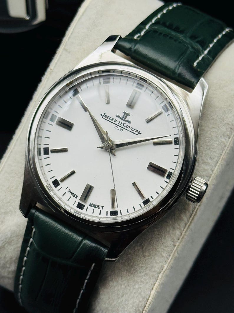 May include: A silver-toned Jaeger-LeCoultre Club watch with a white face and black markers. The watch has silver hands and a green leather strap. The watch face reads "JAEGER-LECOULTRE CLUB" and "SWISS MADE T".