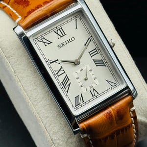Op de afbeelding: Een rechthoekig Seiko horloge met een zilveren kast, witte wijzerplaat en Romeinse cijfers. De band is van bruin leer met witte stiksels. De wijzerplaat toont de merknaam Seiko.