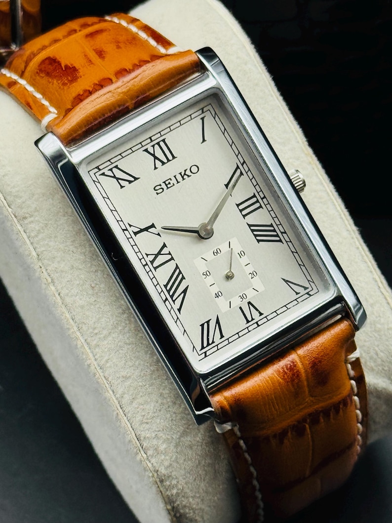 Op de afbeelding: Een rechthoekig Seiko horloge met een zilveren kast, witte wijzerplaat en Romeinse cijfers. De band is van bruin leer met witte stiksels. De wijzerplaat toont de merknaam Seiko.