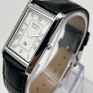 Puede incluir: Reloj Seiko Quartz rectangular con esfera blanca, n&uacute;meros romanos y correa de cuero negro. El reloj tiene un bisel plateado y una peque&ntilde;a corona. La correa tiene una textura con un patr&oacute;n de cocodrilo.