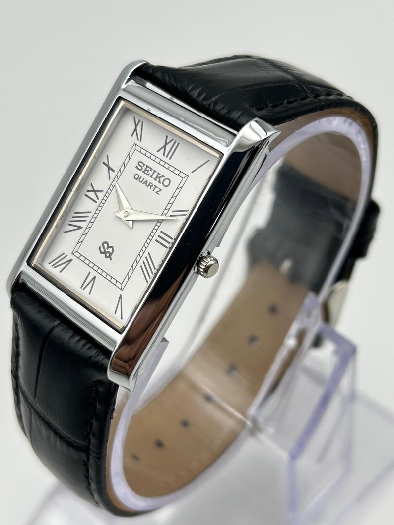 Puede incluir: Reloj Seiko Quartz rectangular con esfera blanca, n&uacute;meros romanos y correa de cuero negro. El reloj tiene un bisel plateado y una peque&ntilde;a corona. La correa tiene una textura con un patr&oacute;n de cocodrilo.
