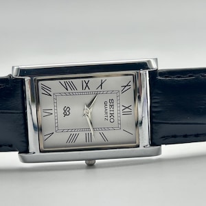 Puede incluir: Un reloj Seiko Quartz rectangular con caja plateada y esfera blanca con n&uacute;meros romanos. El reloj tiene una correa de cuero negro. La esfera del reloj muestra el logotipo de Seiko Quartz.