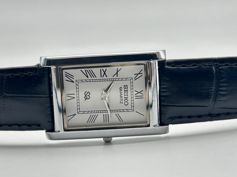 Puede incluir: Un reloj Seiko Quartz rectangular con caja plateada y esfera blanca con n&uacute;meros romanos. El reloj tiene una correa de cuero negro. La esfera del reloj muestra el logotipo de Seiko Quartz.
