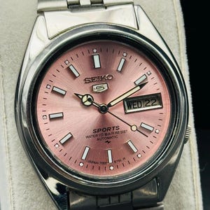 May include: A stainless steel Seiko 5 Sports automatic watch with a pink dial. The watch face displays the day and date, with white rectangular hour markers and hands. The watch band is made of stainless steel.
