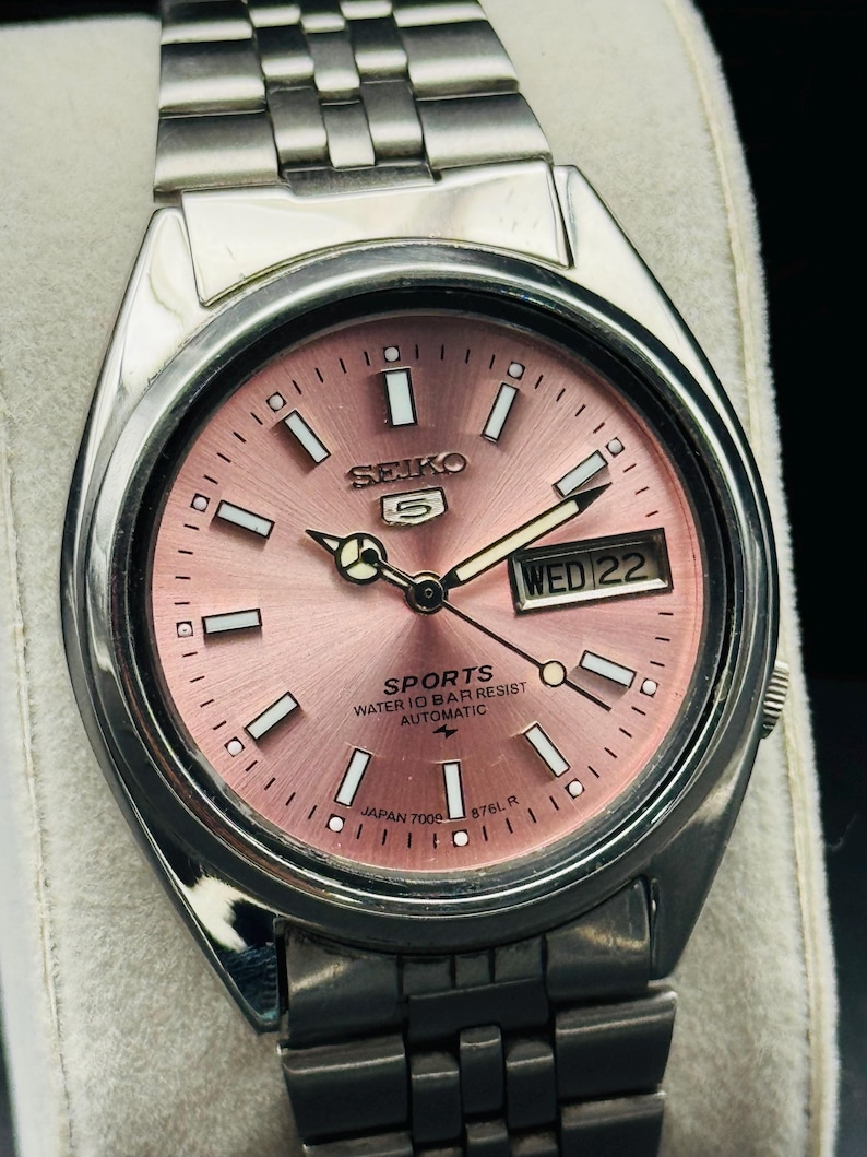 May include: A stainless steel Seiko 5 Sports automatic watch with a pink dial. The watch face displays the day and date, with white rectangular hour markers and hands. The watch band is made of stainless steel.