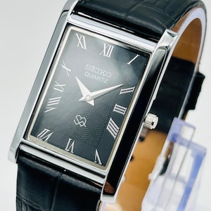 Könnte beinhalten: Rechteckige Seiko Quartz-Uhr mit schwarzem Zifferblatt und silberfarbenem Gehäuse. Die Uhr hat römische Ziffern und ein schwarzes Lederarmband. Das Zifferblatt hat ein strukturiertes Muster und das Seiko-Logo. Eine klassische Uhr.
