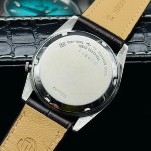 May include: A close-up of a vintage Seiko watch, showcasing the stainless steel back with text including "WATER RESISTANT" and "GENUINE LEATHER" on the brown leather strap. The watch has a silver case and a dark brown leather band.