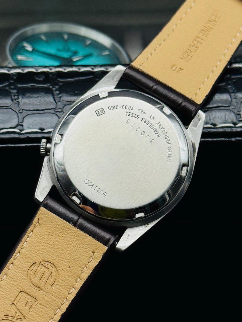May include: A close-up of a vintage Seiko watch, showcasing the stainless steel back with text including "WATER RESISTANT" and "GENUINE LEATHER" on the brown leather strap. The watch has a silver case and a dark brown leather band.