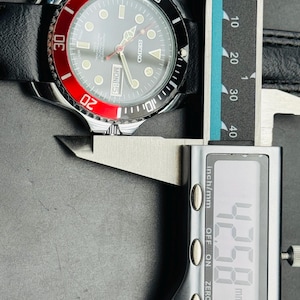 May include: A Seiko watch with a black leather band and a black and red bezel. The watch face is gray with luminous markers and hands. The digital caliper shows a measurement of 42.58 mm.