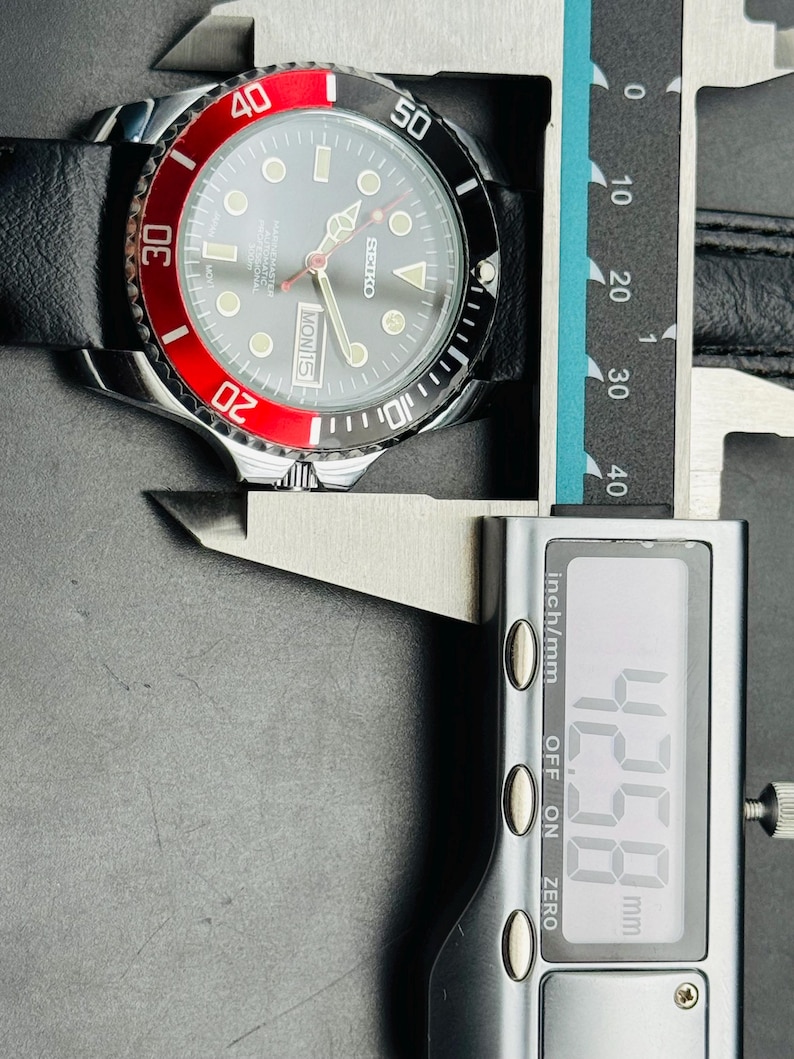 May include: A Seiko watch with a black leather band and a black and red bezel. The watch face is gray with luminous markers and hands. The digital caliper shows a measurement of 42.58 mm.