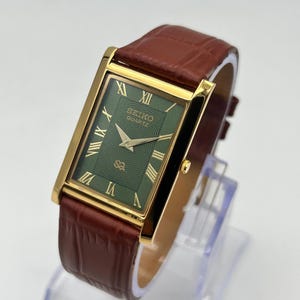 Könnte beinhalten: Eine goldfarbene Seiko Quartz-Uhr mit einem rechteckigen grünen Zifferblatt und goldenen römischen Ziffern. Die Uhr hat ein braunes Lederarmband. Das Zifferblatt trägt das Seiko Quartz-Logo. Die Uhr wird auf einem transparenten Ständer präsentiert.