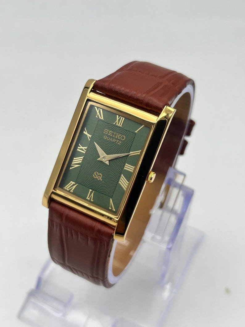 Puede incluir: Un reloj Seiko Quartz dorado con una esfera rectangular verde y n&uacute;meros romanos dorados. El reloj tiene una correa de cuero marr&oacute;n. La esfera del reloj tiene el logotipo de Seiko Quartz. El reloj se exhibe sobre un soporte transparente.
