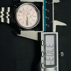 May include: A stainless steel wristwatch with a pink dial is measured with a digital caliper. The watch face displays the day and date. The caliper reads 37.03 mm, approximately 1.46 inches.