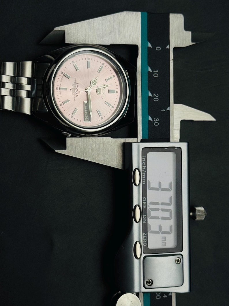 May include: A stainless steel wristwatch with a pink dial is measured with a digital caliper. The watch face displays the day and date. The caliper reads 37.03 mm, approximately 1.46 inches.