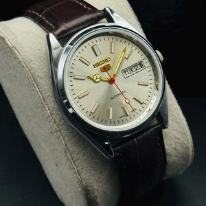 May include: Silver-toned Seiko automatic watch with a light-colored face, gold hands, and a red second hand. The watch features a day and date display and a brown leather strap. The watch is displayed on a gray cushion.
